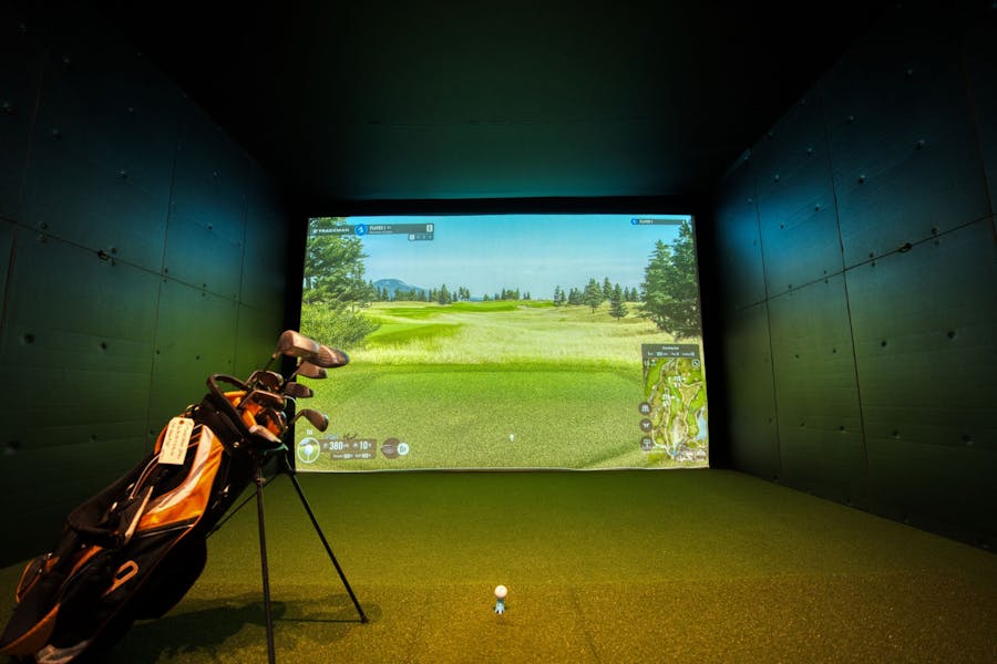 Home golf simulator setup featuring a large projection screen displaying a golf course, with a golf bag and clubs positioned on green turf in front of the screen.