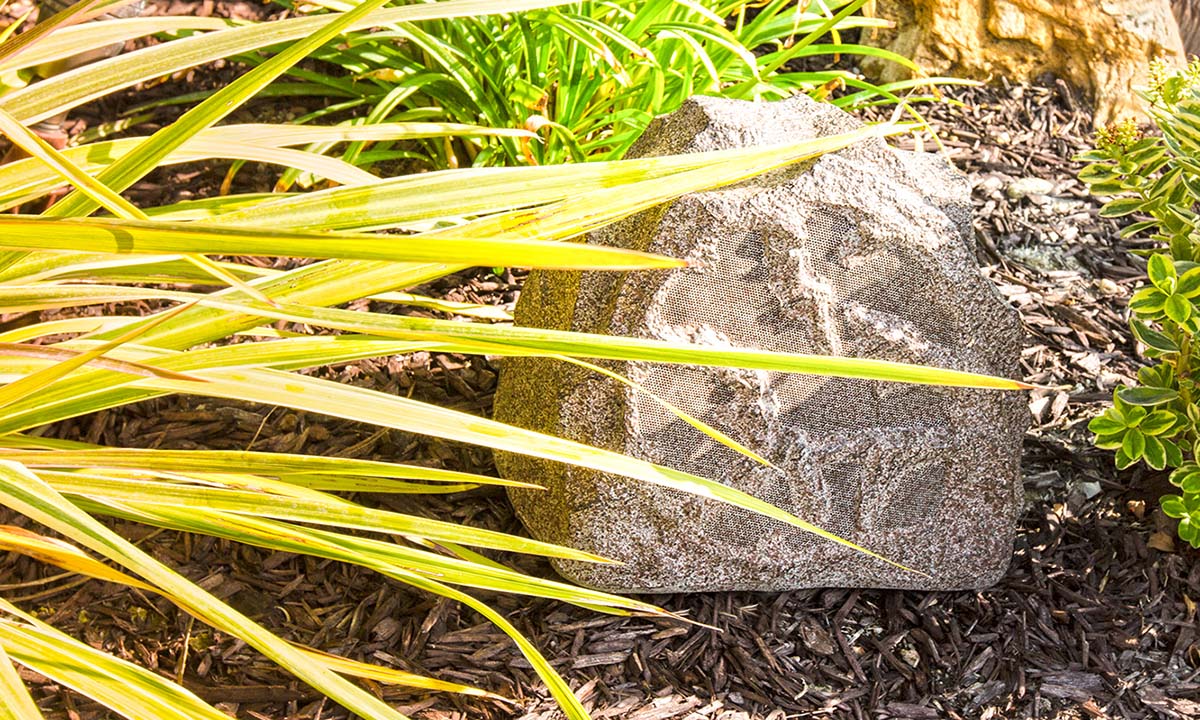 A camouflaged outdoor speaker designed to look like a natural rock, placed among plants and mulch in a landscaped garden setting.