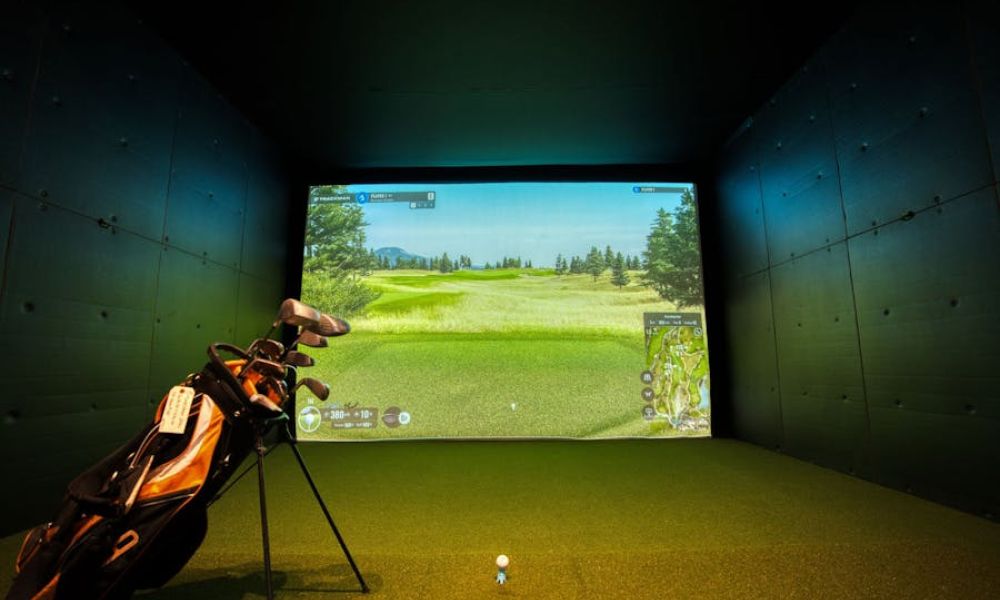 Home golf simulator setup featuring a large projection screen displaying a golf course, with a golf bag and clubs positioned on green turf in front of the screen.