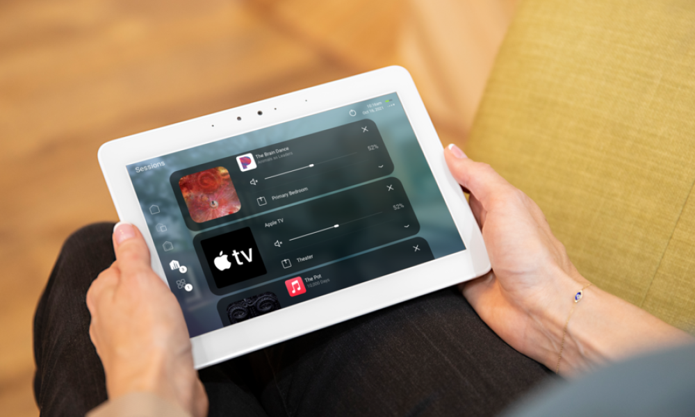 Person holding a tablet with smart home AV control system, managing music and Apple TV across multiple rooms.